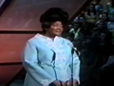 Mahalia Jackson is wearing a blue dress and singing whereas people are watching.